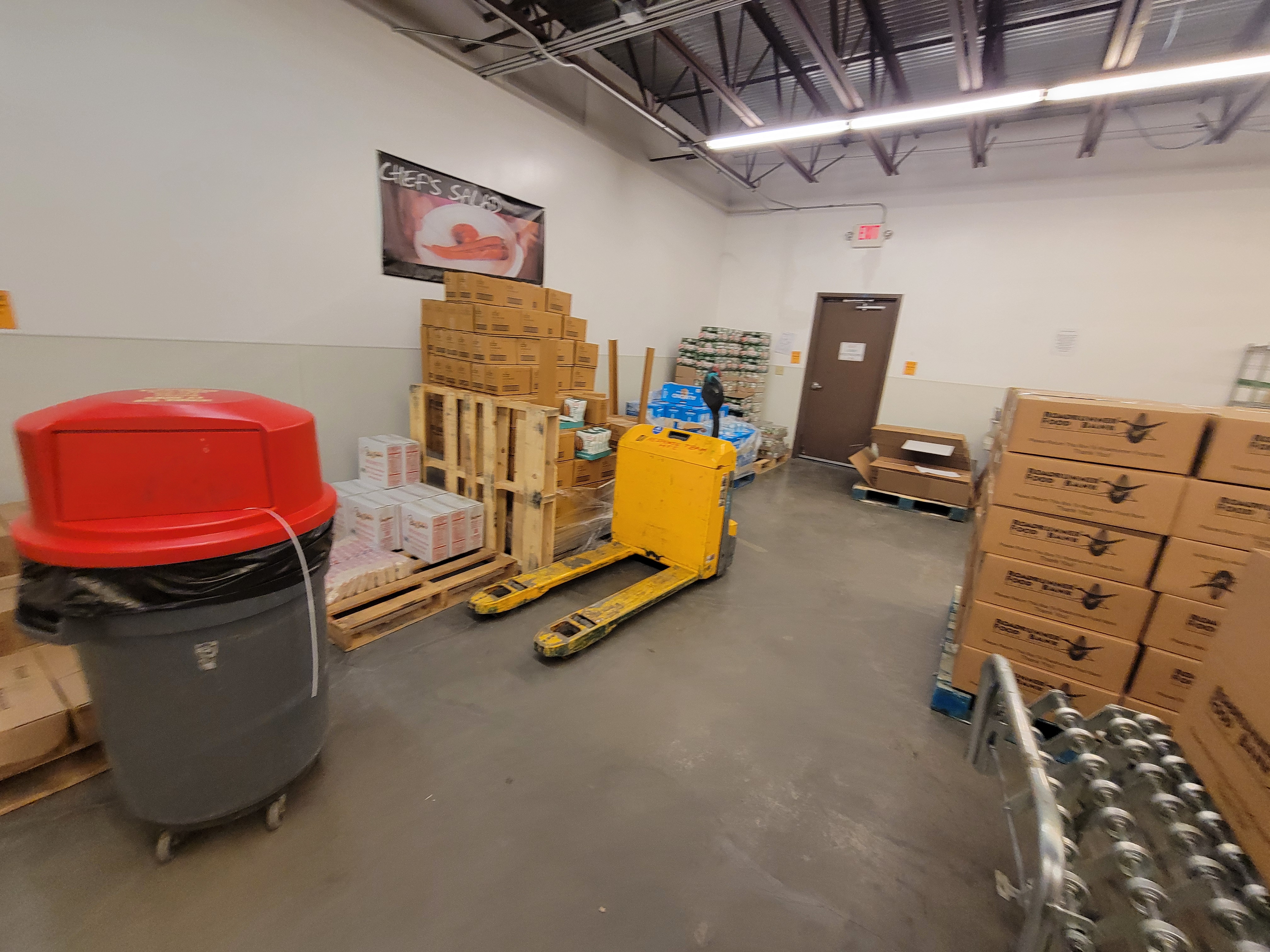 Pallets of food behind a pallet jacket and trash can. Next to tbose are a pallet of boxes and conveyor belt with a box on top.